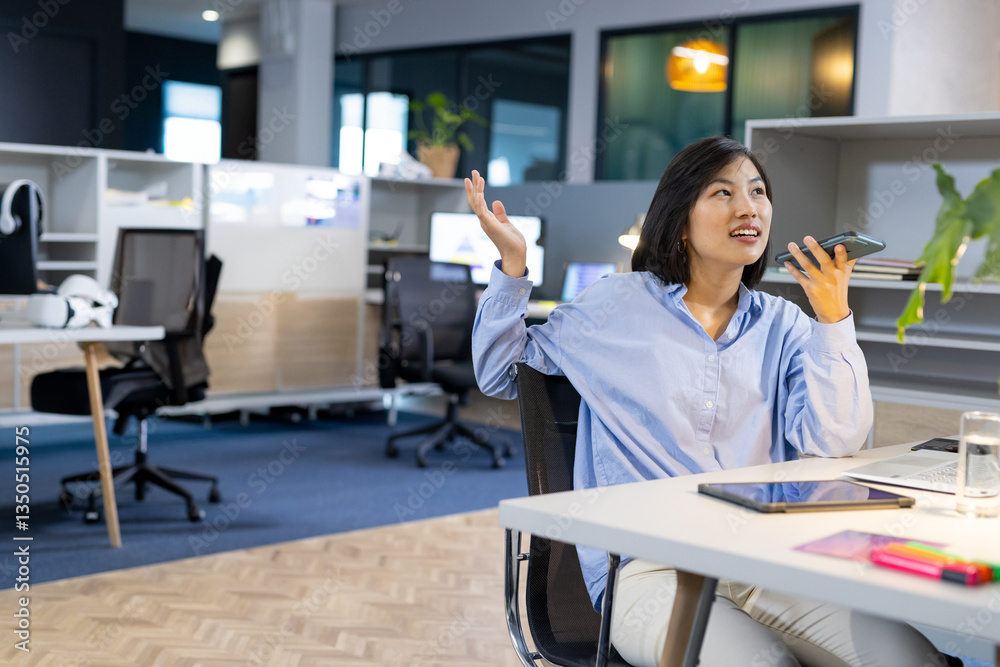 Obraz premium Asian woman in office talking on smartphone, gesturing with hand, looking engaged, copy space