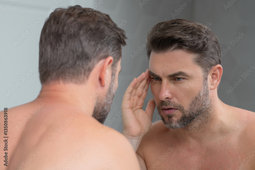 Fototapeta premium Attractive man with perfect skin touch face after shaving. Skin care healthcare cosmetic procedures concept. Close up man looking in mirror, sensitive skin, cosmetology treatment. Skin care.