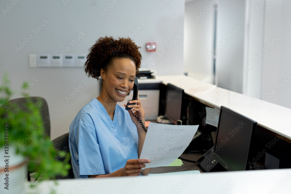 Hospital receptionist in scrubs smiling while talking on phone and holding document Hospital receptionist in scrubs smiling while talking on phone and holding document