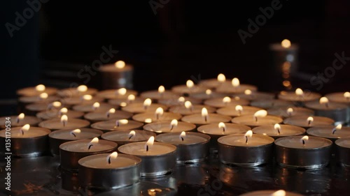 Close-up of burning tealight candles arranged on reflective surface in dark setting. Candlelight and remembrance concept for design and print.