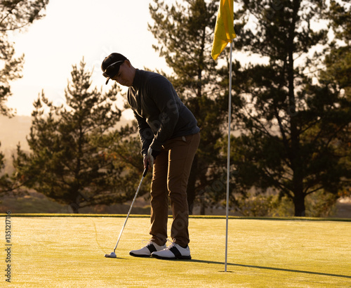 Male golfer focusing on putting ball into hole on sunny golf course