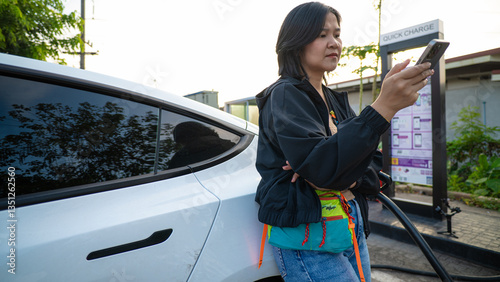 Asian woman tapping on smartphone use electric car is charging outdoor in city at charge station outside