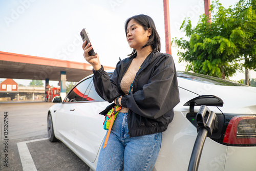 Asian woman tapping on smartphone use electric car is charging outdoor in city at charge station outside
