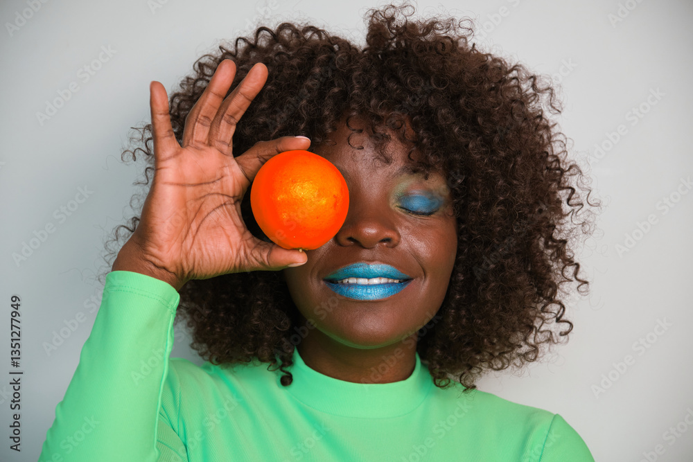 Smiling woman with blue lipstick and eyeshadow holding an orange in front of her eye