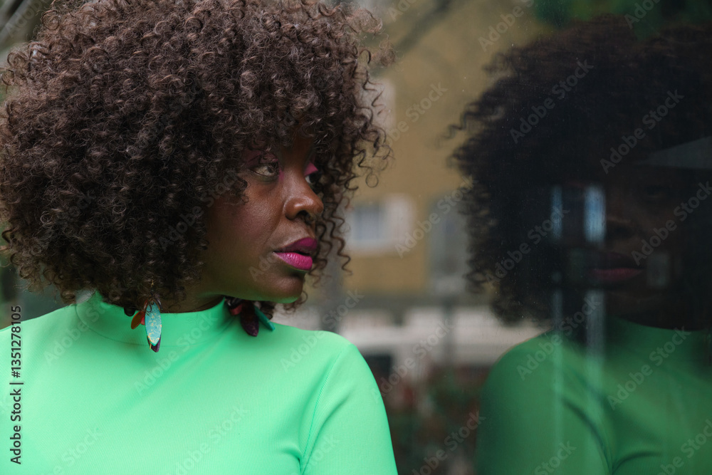 Fashionable black woman with curly hair and colorful earrings looking away and reflecting in a window glass