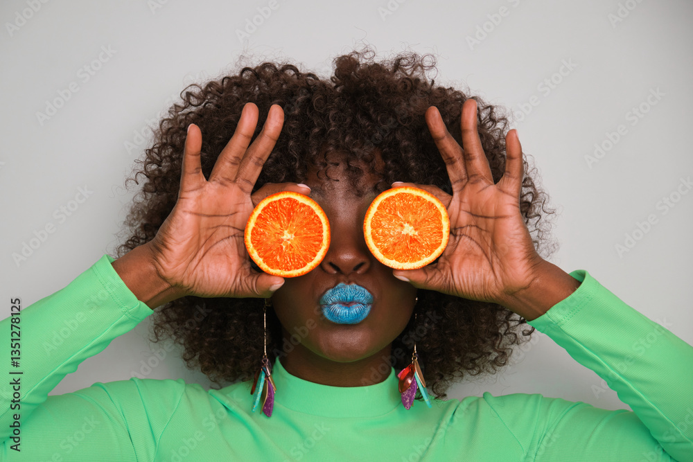 Portrait of a young woman with curly hair holding orange slices covering her eyes and pouting with blue lipstick