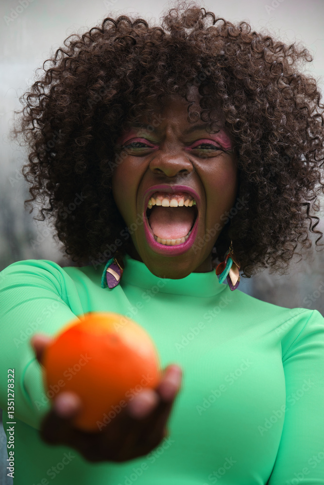 Young model with curly hair and green shirt offering a ripe orange with an angry expression and open mouth