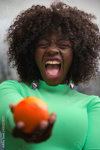 Obraz na płótnie Young model with curly hair and green shirt offering a ripe orange with an angry expression and open mouth w salonie