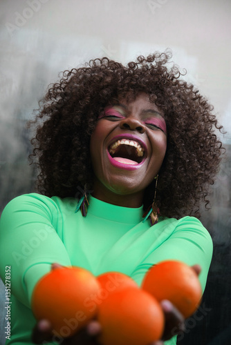 Cheerful woman with curly hair and a bright green shirt laughs heartily while offering fresh oranges