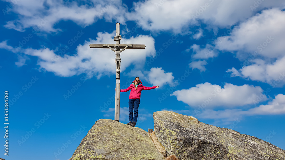 Hiker woman stands with open arms next to a summit cross on Seinerkreuz, Packalpe, Austria, under a partly cloudy sky. Celebrating the peak of the mountain with a scenic view. Remote Austrian Alps
