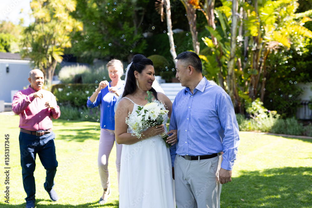 In garden, diverse bride and groom walking while friends celebrating with bubbles In garden, diverse bride and groom walking while friends celebrating with bubbles