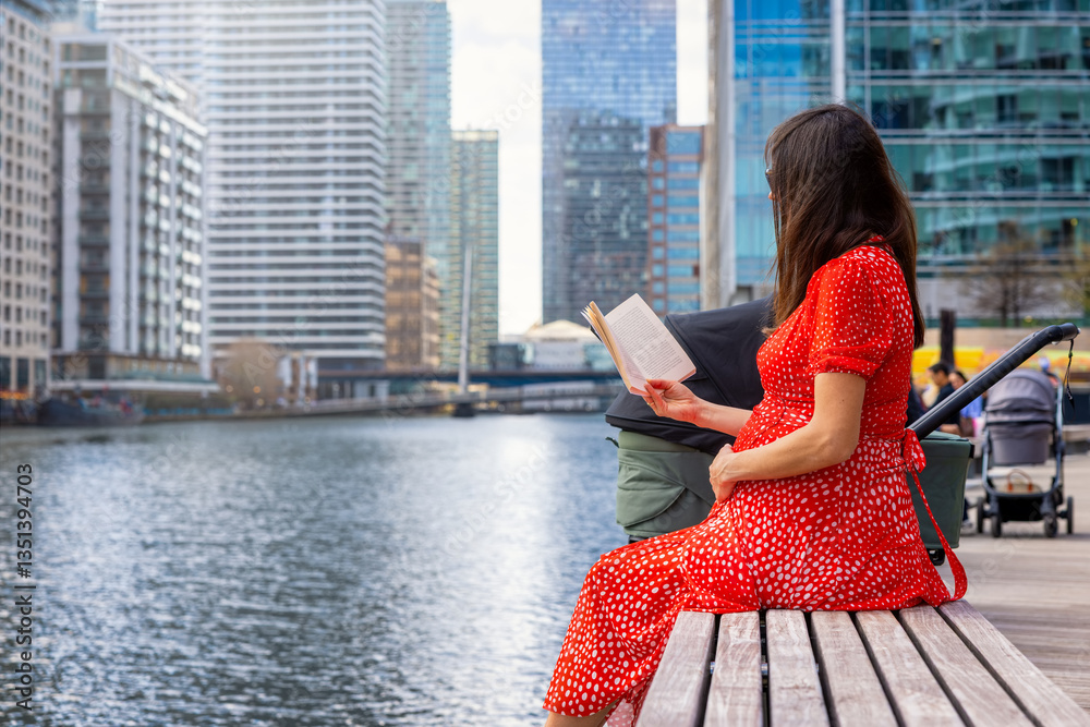 A pregnant mother sits on a bench by the river in a urban city and reads a book
