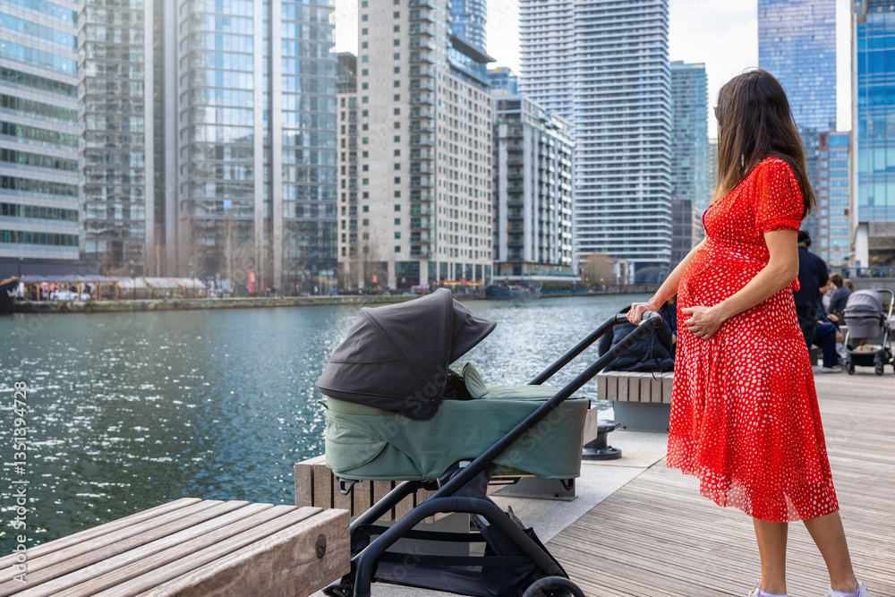 A pregnant mother goes for a walk by the river in a urban city with a baby push chair on a sunny day