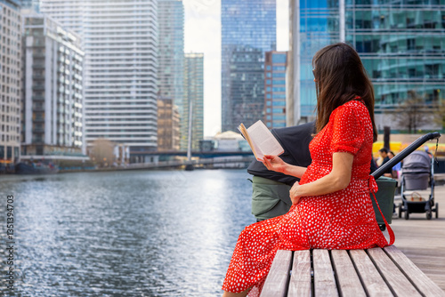 Obraz na płótnie A pregnant mother sits on a bench by the river in a urban city and reads a book w salonie