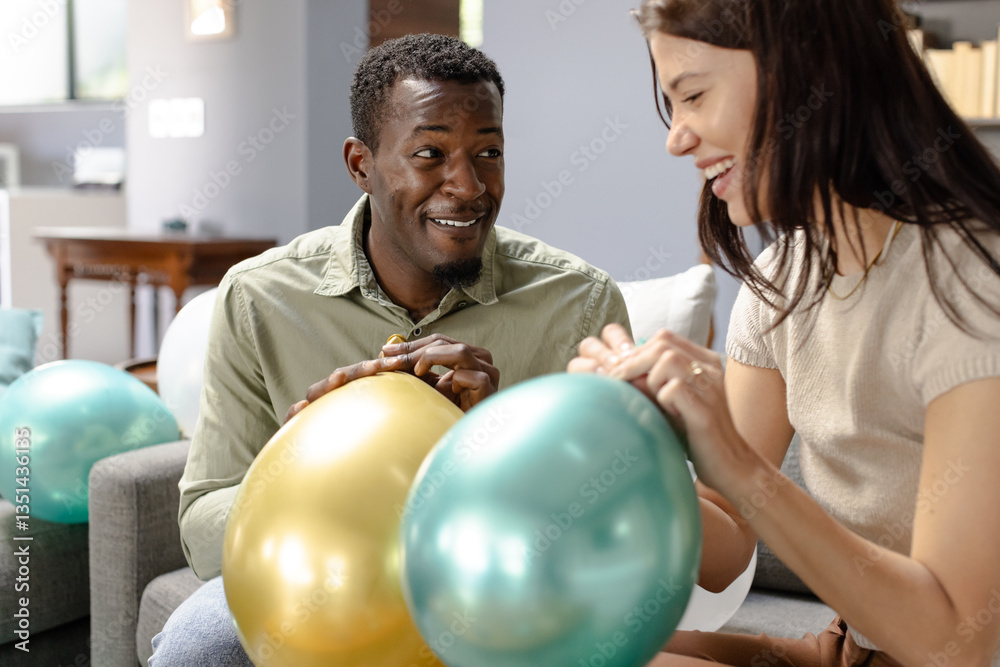 Inflating colorful balloons, Diverse couple smiling during festive preparations at home