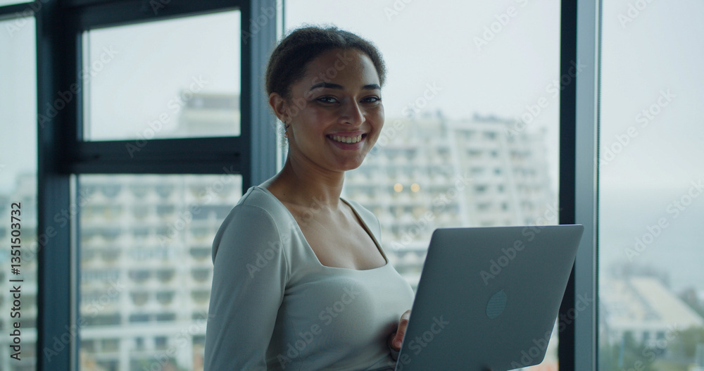 A young professional smiling confidently while holding a laptop near a large window in a modern office with a blurred cityscape in the background A young professional smiling confidently while holding a laptop near a large window in a modern office with a blurred cityscape in the background