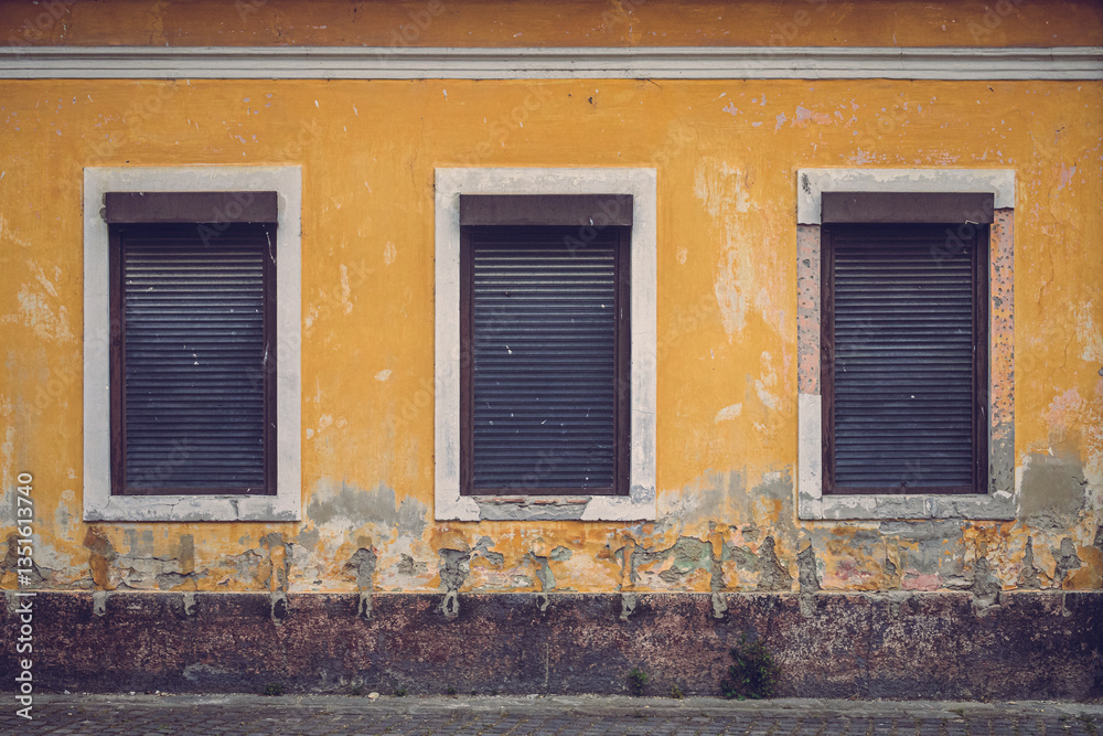 A weathered yellow wall with three closed shutters creates a striking geometric composition. The peeling paint and textured decay evoke a sense of time, history, and forgotten stories A weathered yellow wall with three closed shutters creates a striking geometric composition. The peeling paint and textured decay evoke a sense of time, history, and forgotten stories