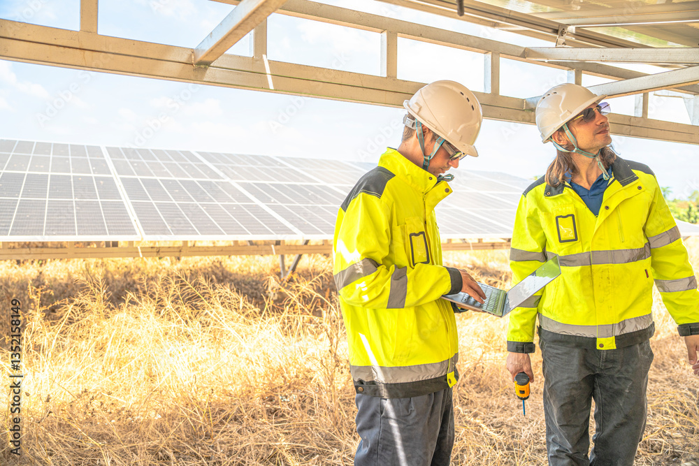 Two solar technicians in bright reflective jackets and helmets are inspecting the wiring under solar panels. One holds laptop for data logging, while the other examines electrical connections closely. Two solar technicians in bright reflective jackets and helmets are inspecting the wiring under solar panels. One holds laptop for data logging, while the other examines electrical connections closely.