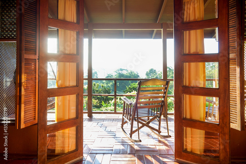 wooden glass doors opening out onto a balcony with a beautiful wooden rocking chair looking out over a tropical garden with bright sun shining onto the balcony, Arenal, Costa Rica, copy space