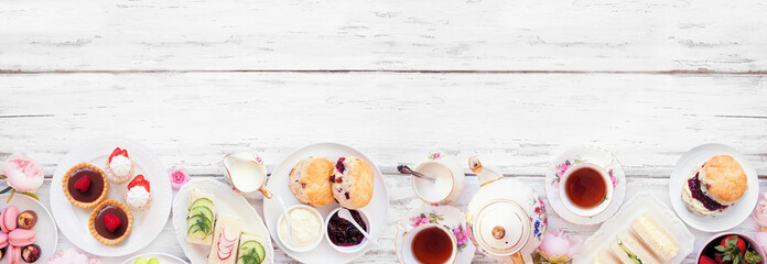Afternoon tea bottom border against a white wood banner background. Vintage tea set, finger sandwiches, scones, and desserts. Top view with copy space.