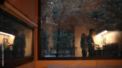 A couple shares an intimate moment, hugging and laughing in the kitchen, with a wintery forest visible through the window. The warm, cozy atmosphere makes it perfect for home and relationship themes.