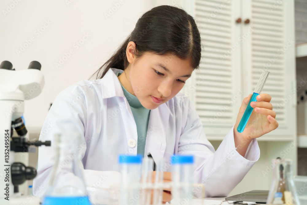 Caucasian teenage girl wearing white lab coat sitting at laboratory desk holding test tube filled with blue liquid while writing on paper during school science experiment session indoors Caucasian teenage girl wearing white lab coat sitting at laboratory desk holding test tube filled with blue liquid while writing on paper during school science experiment session indoors
