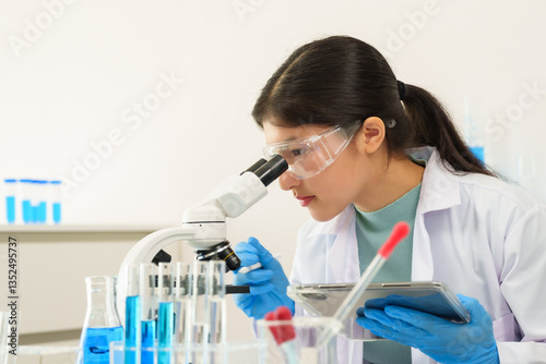 Teenage caucasian girl in lab coat safety goggles and gloves using microscope and holding tablet while performing experiment during high school science lesson at classroom desk