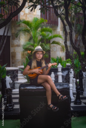 Obraz na płótnie A spirited young girl wearing a trendy hat joyfully strums her ukulele, surrounded by vibrant lush greenery and whimsical chess pieces in a beautiful tropical paradise, Bali, Indonesia w salonie