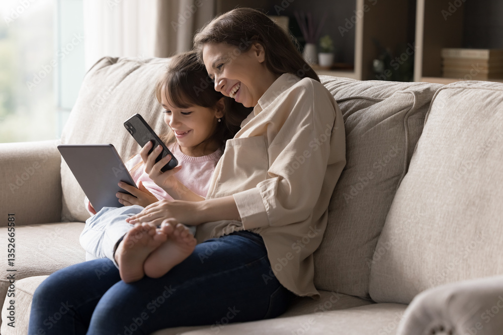 Digital tech and family time. Happy single mom little child daughter cuddle on couch at home enjoy using gadgets sharing data via wifi playing online game together from diverse accounts. Copy space Digital tech and family time. Happy single mom little child daughter cuddle on couch at home enjoy using gadgets sharing data via wifi playing online game together from diverse accounts. Copy space