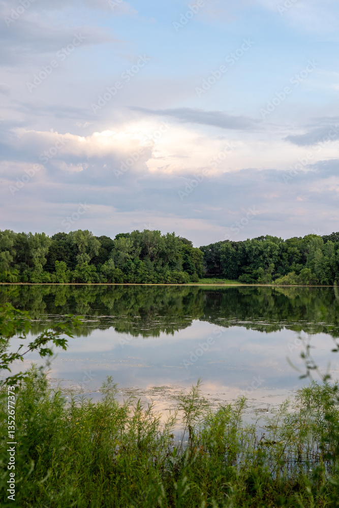 Obraz premium Still lake reflecting the sky and trees, framed by green foliage.