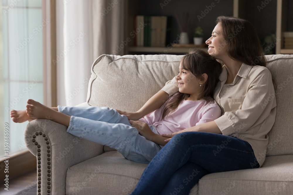 Serene dreams and togetherness. Affectionate millennial mother sitting on sofa holding beloved little daughter in arms looking away talking to kid with tender smile dreaming imagining future together