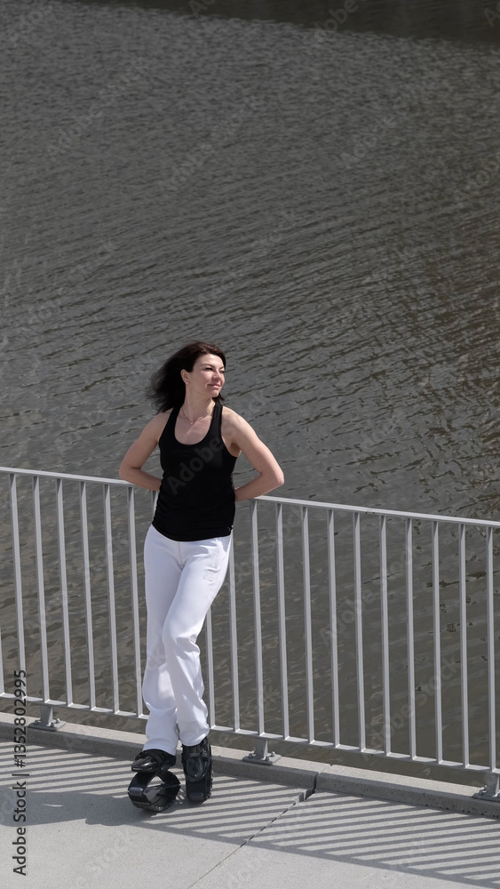 Fototapeta premium A woman jumps energetically on kangaroo boots by the riverside, embracing outdoor fitness with a smile. The blend of innovative exercise gear and urban scenery showcases the evolution of modern