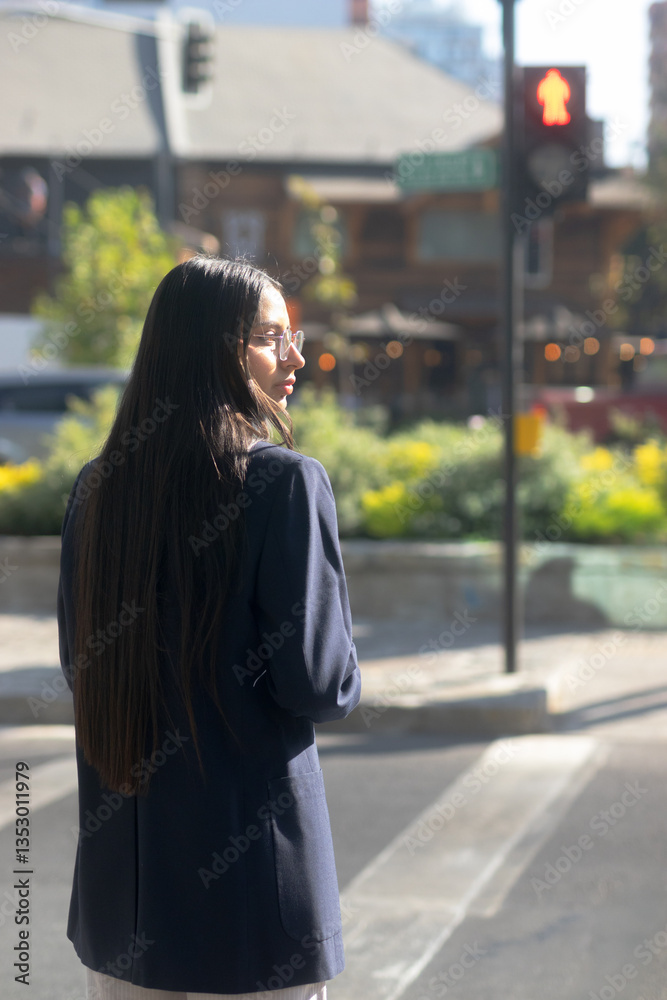 Fototapeta premium Young Hispanic businesswoman with blue blazer, white shirt, looking at stopped cars – Red traffic light in the background