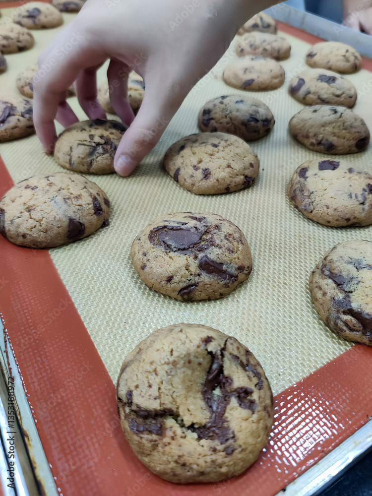 Chocolate chip cookies cool on a baking tray while someone reaches for a warm treat in a cozy kitchen setting Chocolate chip cookies cool on a baking tray while someone reaches for a warm treat in a cozy kitchen setting