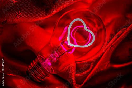 A creative and artistic macro photograph of a light bulb with a glowing filament shaped like a heart. The image is captured with red lighting, creating a warm, passionate, and romantic atmosphere. 