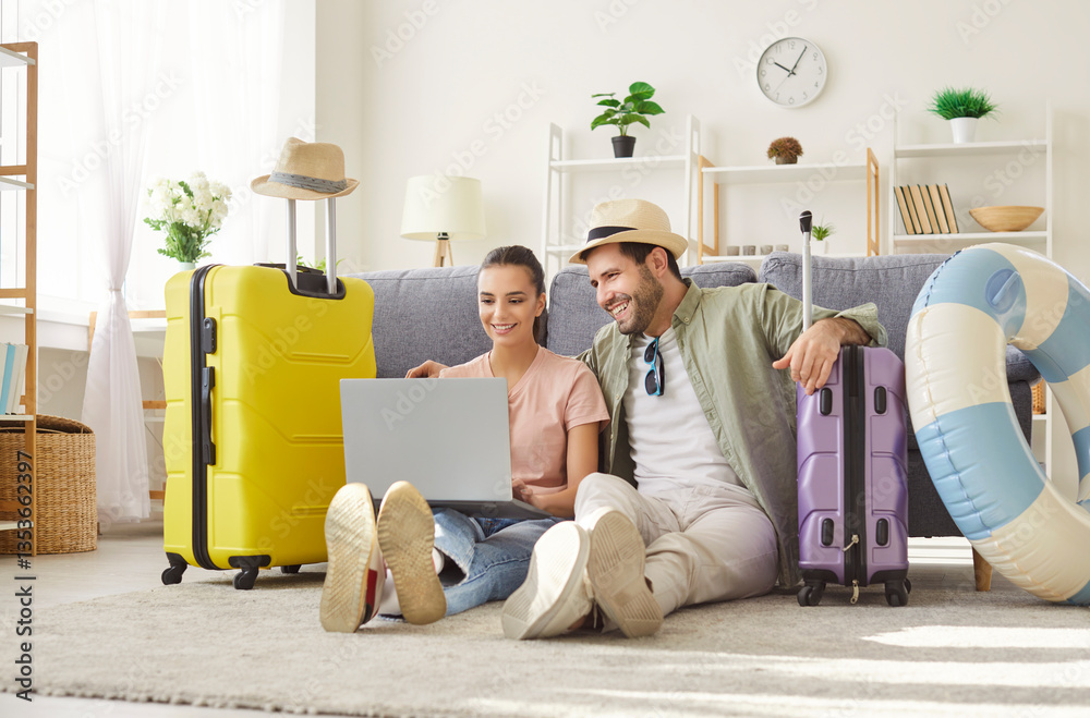 Fototapeta premium Happy smiling couple travelers sitting on floor with suitcases at home booking tickets online via laptop. Young man and woman wearing beach hat choosing tour for holiday trip. Vacation concept.