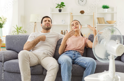 Young couple being exhausted of summer heat, using electric fan to make some fresh air, sitting on couch and waving hands, sweating and feeling very hot. What a heat, we cannot stand