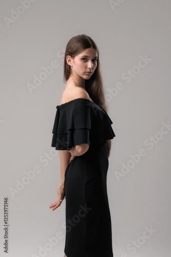 Young woman poses elegantly in a stylish black off-shoulder dress against a neutral background
