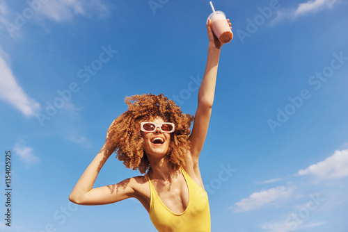 Joyful woman enjoying a summer day on the beach with a smoothie, wearing stylish sunglasses against a bright blue sky