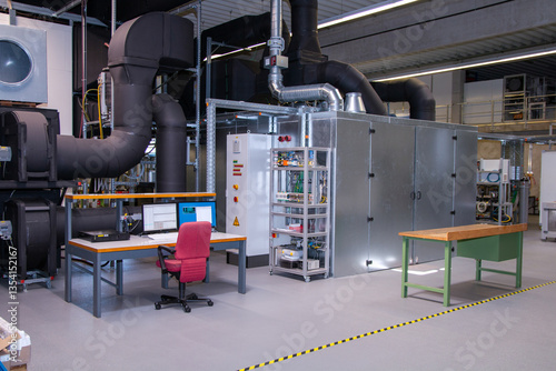 Full view of industrial HVAC testing station with insulated air ducts, control cabinets, engineering workstation with computer monitors and server, and empty workbench