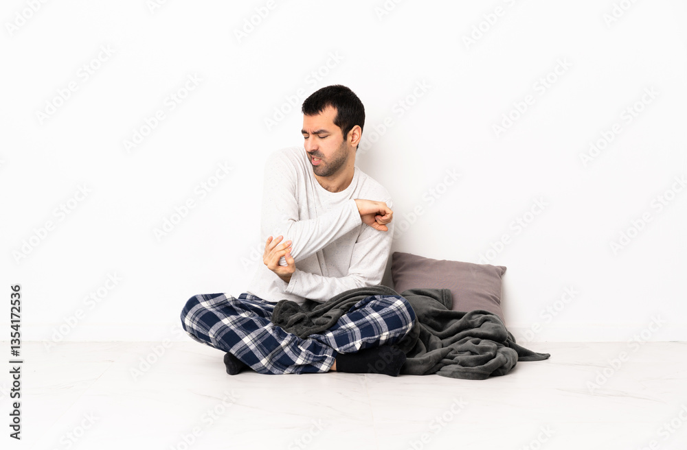 Caucasian man in pajamas sitting on the floor at indoors with pain in elbow Caucasian man in pajamas sitting on the floor at indoors with pain in elbow
