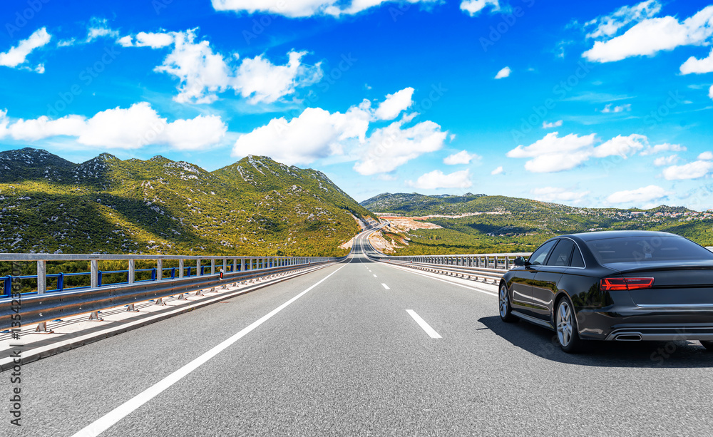 A sleek black luxury sedan drives along a scenic highway surrounded by lush green mountains under a bright blue sky with fluffy clouds.