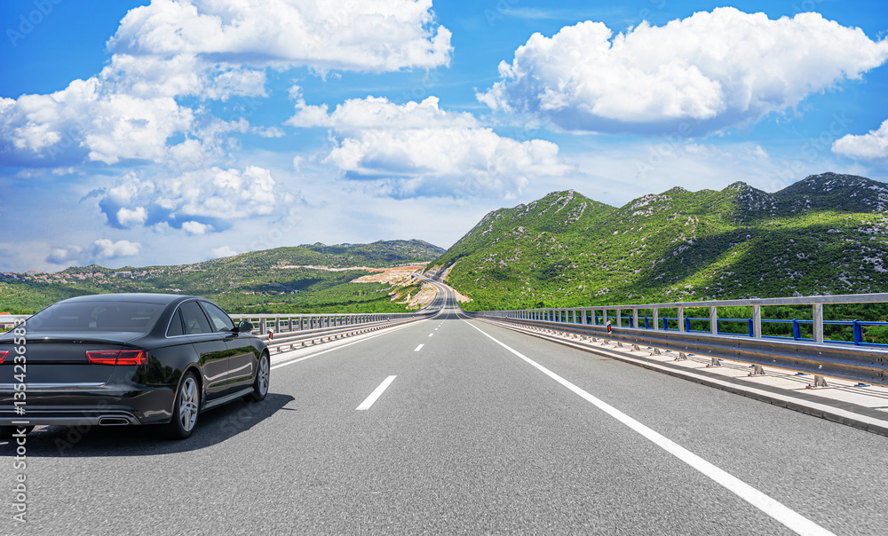 A black luxury sedan cruises along a well-maintained highway surrounded by lush green mountains under a bright blue sky filled with fluffy white clouds.