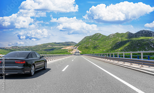 Obraz na płótnie A black luxury sedan cruises along a well-maintained highway surrounded by lush green mountains under a bright blue sky filled with fluffy white clouds. w salonie