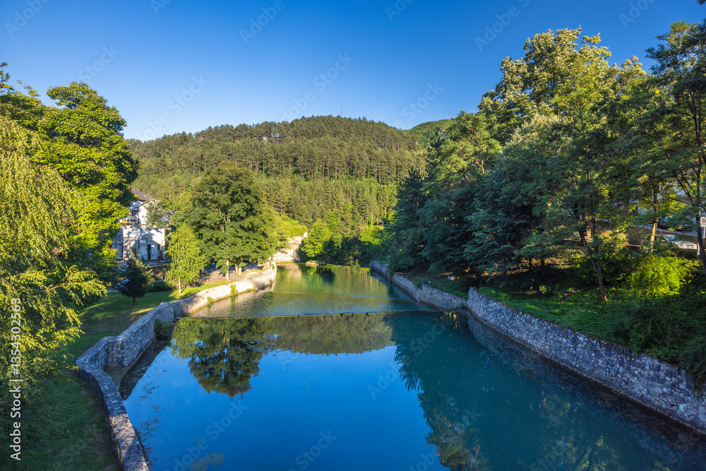 Fototapeta premium The Pliva river in Jajce town in Bosnia and Herzegovina, Europe. Scenic river view with stone embankments surrounded by lush green trees and a forested hillside under a clear blue sky.