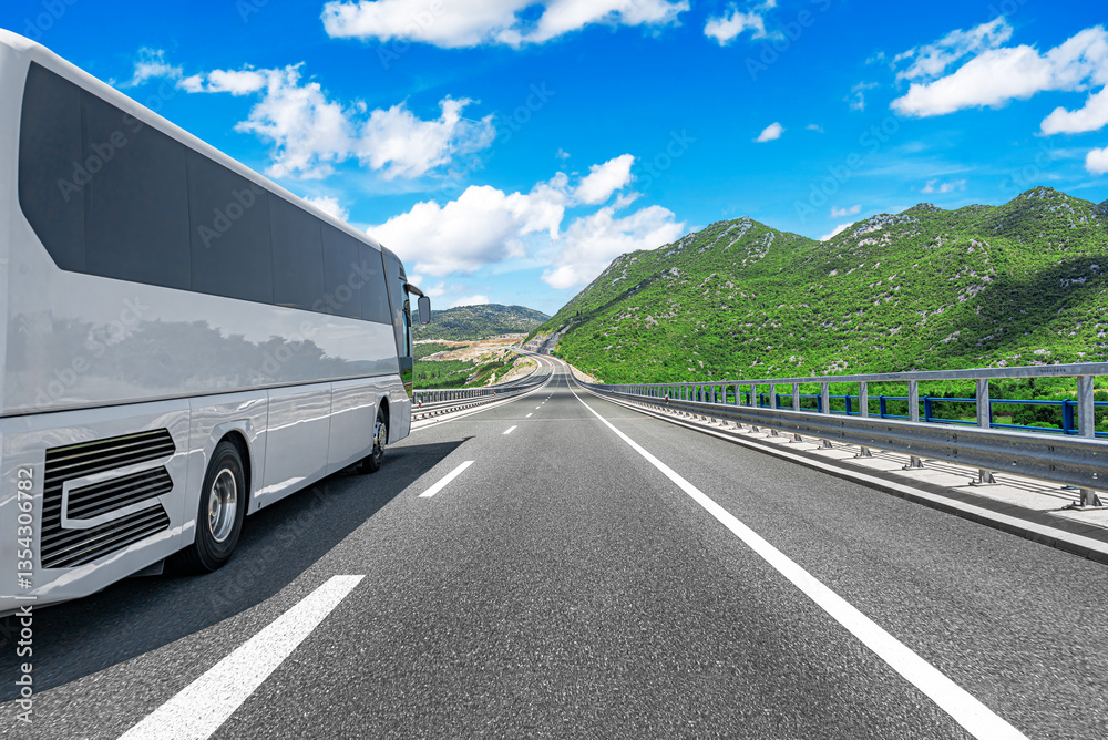 A modern white coach bus travels along a smooth highway surrounded by lush green mountains under a bright blue sky with scattered clouds.