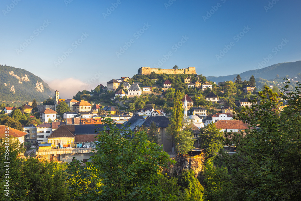 Fototapeta premium Jajce town in Bosnia and Herzegovina, Europe. Picturesque view of a European town nestled on a hillside with a historic fortress overlooking the houses, captured under a clear blue sky.