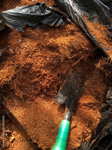 Top view of loose cocopeat, also known as coconut coir, in a black plastic bag with a small green-handled gardening trowel placed on top. Rich brown texture of natural fibers, commonly used for soil 