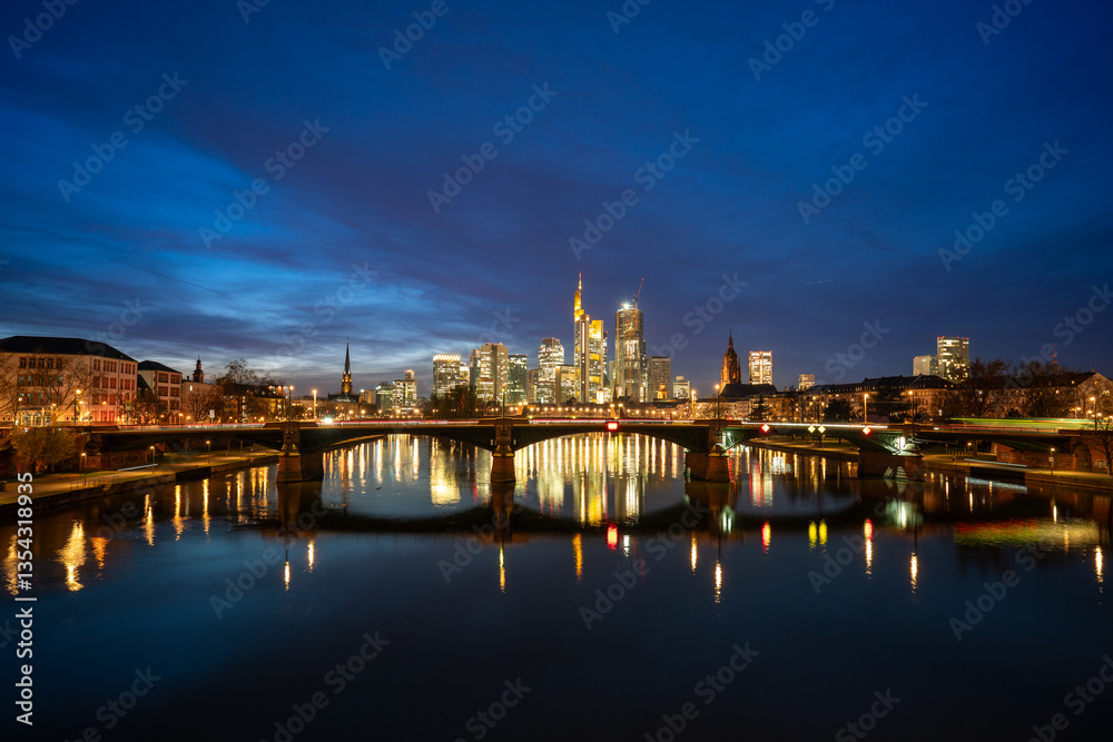 Frankfurt cityscape at night, with illuminated skyscrapers reflecting on the Main River, a lit bridge, and a clear dark blue sky, showcasing urban beauty and tranquility.