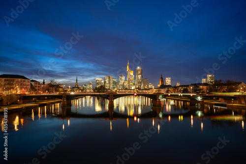 Frankfurt cityscape at night, with illuminated skyscrapers reflecting on the Main River, a lit bridge, and a clear dark blue sky, showcasing urban beauty and tranquility.